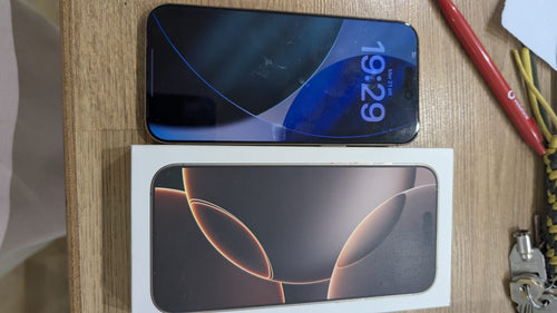 iPhone 16 Pro Max usato con lo schermo acceso, appoggiato accanto alla sua scatola originale su un tavolo di legno